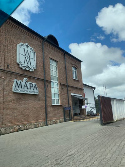 This is a daytime, eye-level, outdoor photograph of the exterior of a building. The building is constructed of red brick and has a stone foundation. A prominent white sign with a stylized "WM" emblem is mounted on the brick facade. Below this, a larger, 3D sign reads "MAPA" in large letters, with "Мастацкая галерэя" in smaller letters below it, indicating it is an art gallery. The building has several tall, multi-paned windows. Adjacent to the main building is a white structure with a sloped roof. A metal gate with signs, including one that reads "ОТКРЫТО" (Open), is visible. A rusty corrugated metal fence extends to the right of the gate. The ground in front of the building is paved with interlocking concrete blocks. The sky is blue with scattered white clouds. The location is confirmed to be in Nesvizh, Belarus.