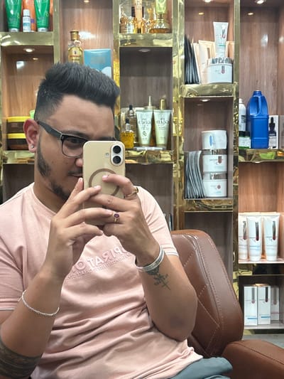 The image captures a male individual taking a mirror selfie inside what appears to be a salon or barbershop in South Dumdum, India. The subject, with dark hair styled upwards and faded sides, a light beard, and black-rimmed glasses, is wearing a light pink polo shirt and light-colored pants. He is seated in a brown, diamond-stitched upholstered chair. He wears a silver chain bracelet on his left wrist and a silver-colored bangle on his right wrist. A plain gold-colored ring is on his right ring finger, and a gold-colored ring with a prominent red stone is on his right middle finger. A tattoo featuring a cross and a rose design is visible on his outer right forearm, with another partial tattoo on his inner left forearm. He holds a light-colored smartphone with dual rear cameras and a visible MagSafe ring in both hands, angling it towards his reflection in a mirror to take a photograph. The background consists of display shelves made of wood with gold-colored metallic framing, illuminated by integrated lighting. These shelves are stocked with various cosmetic and grooming products, including tubes, bottles, jars, and a blue plastic container.