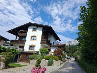 In the city of Münster, Austria, a residential street curves gently uphill. The street is paved with asphalt and bordered by a lush green hedge on the right. On the left, a multi-story white house with dark brown trim and balconies dominates the foreground. The house features multiple windows with ornate trim and a steeply pitched roof. In front of the house, a tiered garden beds filled with various plants and flowers, including vibrant pink petunias in a pot, are visible. A wooden garage with an open door is attached to the side of the house. Further up the street, a dark blue car is parked. The background reveals a dense forest and a majestic mountain range under a bright blue sky dotted with fluffy white clouds.
