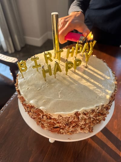 The scene depicts a person lighting gold "HAPPY BIRTHDAY" candles on a cake with white frosting and a pecan-covered edge. The person is using a pink lighter and a gold wand to light the candles. The cake sits on a white plate. The setting appears to be indoors, with a wooden table visible and shadows suggesting natural light. There is no indication of the location being Santiago Yancuitlalpan, Mexico.