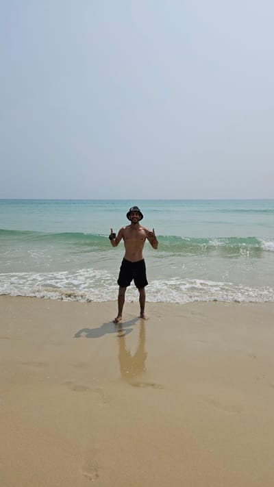 A man is captured in a full-body shot, posing on a sunny beach at the edge of the ocean in Bac Cam Ranh, Vietnam. He stands shirtless, wearing black shorts and a dark bucket hat. With a wide smile, he gestures with both hands, appearing happy and relaxed.

The man is positioned where gentle waves break onto the shore, creating a white foam line against the clear blue-green water. The ocean extends to a light blue horizon under a bright, slightly hazy sky. To the man's right, golden-brown sand stretches along the beach, marked by his shadow cast towards the left and several footprints in the wet shore. The scene is illuminated by bright daylight, indicating clear and pleasant weather conditions. No visible text is present in the image.