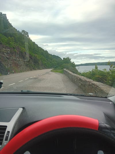 A view from inside a vehicle on a road in Dores, United Kingdom, shows a scenic landscape under an overcast sky. The road curves to the left, bordered by a steep, rocky hillside covered in lush green vegetation and some scattered trees. To the right of the road, a low stone wall separates the road from a body of water, likely Loch Ness, which stretches into the distance. The water is calm and reflects the muted sky. The interior of the vehicle is visible in the foreground, with the dashboard and a red steering wheel cover clearly in view. There are no people visible in the image. The scene appears to be a typical day for driving along the shores of Loch Ness.