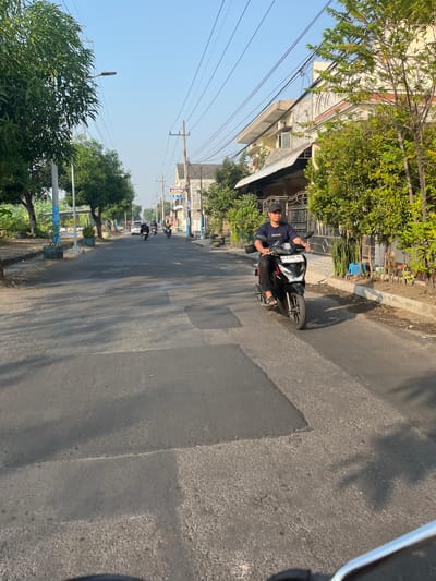 A young man wearing a dark blue t-shirt and a black baseball cap rides a black motorcycle down a paved road in Sugih Waras, Indonesia. He is looking forward and appears to be traveling away from the camera. The road is lined with trees and houses on both sides. The sky is clear and blue, and the sun is shining. There are power lines running along the street. In the background, other motorcycles and a car can be seen. The motorcycle has a license plate that reads "W 5306 SG". The scene depicts a typical day on a residential street in a town in Indonesia.