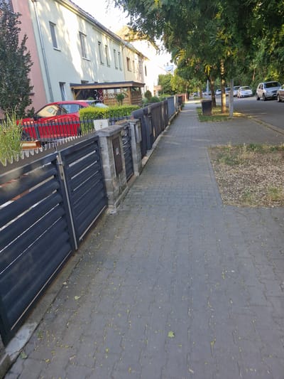 The image shows a residential street in Brno, Czechia. A paved sidewalk runs along the left side of the street, bordered by a dark metal fence and a row of houses. The houses are two to three stories tall and appear to be attached, with light-colored facades. A red car is parked in front of one of the houses. The street itself is paved, with several cars parked on the right side. Trees line the right side of the street, casting shadows on the sidewalk and road. The sky is visible through the trees and appears to be clear and sunny. The scene is calm and quiet, with no people or animals visible.