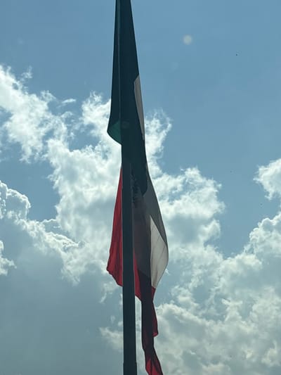 A flag with vertical stripes of green, white, and red flies against a blue sky filled with puffy white clouds. The flag is mounted on a pole and appears to be waving slightly in the wind. The green portion is on the left, followed by white in the center, and red on the right. The flag's design, combined with the sky background, creates a vibrant image.