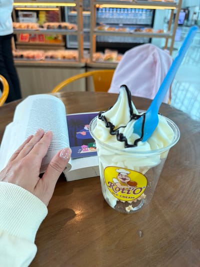 The image captures a close-up view of a wooden table with various items on it. In the foreground, a person's left hand, with white polka dot nail art, rests on an open book. To the right of the book, a clear plastic cup filled with soft-serve ice cream, topped with chocolate sauce, sits on the table. A blue plastic spoon is inserted into the ice cream. The cup has a yellow circular logo with the text "Roti'O ICE CREAM" and a cartoon chef. In the background, a blurred display of baked goods and a pink fabric item are visible, suggesting the setting might be a cafe or bakery. The scene is indoors, and the lighting suggests it is daytime. The location in the city of Rawa Burung, Indonesia is not directly evident from the image itself.