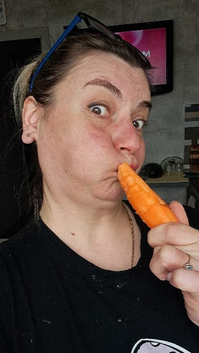 A medium close-up image depicts an adult female positioned indoors in Bel'kovo, Russia. She is looking directly forward with wide-open, brown eyes. Her lips are pursed and pushed outward around the tip of an orange carrot, which she holds with her right hand. Dark-rimmed eyeglasses with blue arms are perched on her head, above her hairline. She wears a dark t-shirt, a thin gold-colored chain necklace, and a ring with a light-colored stone on the fourth finger of her right hand. The background consists of a dark gray, textured wall. To the upper right, a television screen with a dark bezel displays a pink and magenta image with a partial white letter "M." Indistinct objects, possibly glass containers, are visible on a surface to the right.