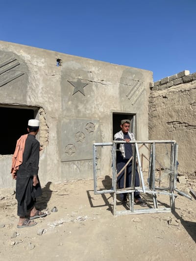 An outdoor scene in Spin Boldak, Afghanistan, depicts two males in an unpaved, dusty area in front of a partially constructed or damaged building under a clear blue sky.
The central structure is a light grey concrete or mud-brick building featuring decorative reliefs: a prominent five-pointed star and three diagonal parallel lines on the upper left, and a lower rectangular panel with three circular designs each containing an internal star. Visible cracks traverse the facade, particularly above the star, and a small, irregular hole is present near the top center of the wall. Two large, empty rectangular openings, consistent with uninstalled windows or doors, are present in this structure. To the right of the main building, a dilapidated mud-brick wall, possibly part of an older or different structure, is visible.
A male individual is positioned within the central doorway of the main building, emerging outwards. He wears a dark long-sleeved tunic (shalwar kameez), dark pants, a light-colored checkered scarf draped over his shoulders, and a patterned cap. He holds a collection of dismantled metal window frames, many with shattered glass remnants, stacked vertically in front of him.
To the left, with his back partially to the viewer, stands another male individual. He wears a dark long-sleeved tunic, dark sandals, and a white cap. An orange-colored fabric is draped over his left shoulder. He faces the central building, with no direct interaction with the other individual visible.
The ground is dry, unpaved, and covered with dirt and construction debris. A small, silver-colored metallic object, possibly a bowl or cup, lies on the ground between the two men. Strong sunlight from the upper right casts distinct shadows across the scene.