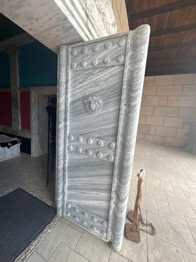 The image shows a large, ornate marble door set in an entryway. The door is carved with circular decorative elements and a central circular relief of a Medusa head. To the left of the door, a dark grey mat is placed on a cobblestone floor. Beyond the door, a passageway with marble walls and red and teal colored sections is visible, with a black metal stand holding a lantern. The background consists of a wooden ceiling and a brick wall. A small wooden stand with a rope is positioned to the right of the marble door. The scene appears to be an exterior view of a building with architectural details suggestive of classical or ancient design. The location is identified as Neofit Rilski, Bulgaria.