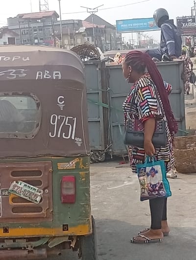 An outdoor shot reveals a woman standing near a parked tricycle. The tricycle is marked with "TOPU," "ABA," and the number "9751." The woman, dressed in a patterned top and leggings, carries a handbag and a blue shopping bag. A man in a helmet sits atop a dumpster in the background. Buildings and signage, including one that says "XASHIN," fill the distant background. The scene is bathed in daylight.