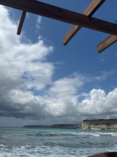 The scene depicts a coastal landscape viewed from a low vantage point, presumably from under a wooden pergola or similar overhead structure, in Episkopi Lemesou, United Kingdom. In the immediate foreground, two intersecting dark brown wooden beams are visible across the top of the frame, partially obscuring the sky. The bottom right corner reveals a portion of a dark-skinned forearm. The midground features the sea, characterized by turquoise water with white-capped waves breaking towards the viewer. In the background, to the right, a series of light-colored cliffs rise abruptly from the sea, topped with sparse, dark green vegetation. Further left, a distant, darker landmass extends along the horizon. The sky overhead is partly cloudy, with clear blue sky visible in the upper right quadrant, transitioning to a mix of white cumulus clouds and darker, heavier clouds towards the left and lower portions of the sky. No other individuals or specific actions are discernible. The environment suggests a Mediterranean climate.