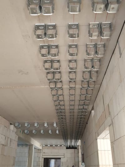 The image shows a ceiling in what appears to be a construction site. Numerous rows of metal brackets are attached to the concrete ceiling. These brackets are spaced evenly and are connected by thin red wires, suggesting they are part of a suspension system for lighting, electrical conduits, or other building services. The overall environment is unfinished, with exposed concrete and block walls visible in the background. There are no people or animals depicted in the image. The location is identified as Osterwieck, Germany, likely a building under construction or renovation.