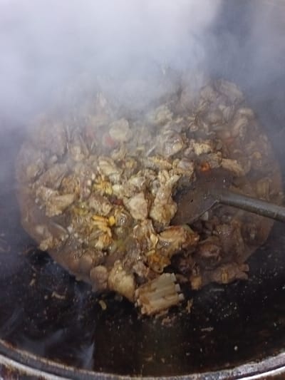 A large, dark, circular cooking vessel, consistent with a kazan, contains a substantial quantity of meat and other ingredients actively cooking. Significant steam rises from the surface of the food, obscuring the upper portion of the scene. The food consists primarily of irregularly shaped pieces of light to dark brown meat, intermixed with smaller fragments of yellow, white, and red components, possibly vegetables such as onions, carrots, and peppers. A dark-colored, broad-bladed utensil, likely a skimmer or spatula, with a visible handle extending towards the upper right, is partially submerged in the food. The liquid base in which the food is cooking appears rich and dark brown. The lower interior and rim of the cooking vessel are visible, showcasing a dark, possibly cast-iron, surface. The scene is located in Kasri Arifan, Uzbekistan. No individuals are visible.