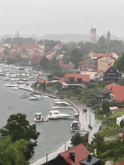 The image depicts a hazy, overcast day in the city of Mikołajki, Poland, as indicated by the faint outline of a tower visible in the distance. The foreground is dominated by a marina filled with numerous boats of varying sizes, docked and seemingly at rest. The water surface appears choppy, reflecting the moody atmosphere. Beyond the marina, the cityscape unfolds with a cluster of buildings characterized by red-tiled roofs. Some of these structures appear to be residential homes, while others, like the taller building with multiple windows and the word "HOTEL" visible, suggest commercial establishments. Lush green trees are interspersed throughout the urban landscape, adding a touch of nature to the scene. The overall impression is one of a tranquil, perhaps even slightly somber, day in a waterfront town.