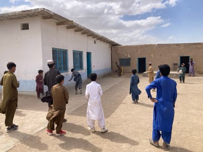 The image captures a scene of young boys playing in the courtyard of a school in Saranan, Pakistan. The location is determined using the coordinates provided (latitude: 30.532, longitude: 66.909). The courtyard is surrounded by a simple, white-washed building on one side, with a larger, earthen-colored building facing the school building, forming an open space. The courtyard is relatively bare, with a dirt floor, and is shaded by a partially cloudy sky.

Nine boys are present, ranging in age from pre-teen to early teens. The boys are engaged in various activities. Some stand and watch, while others are actively playing.  One boy is running toward the right, while another is leaping through the air. One boy appears to be holding a stick, possibly for play. Several of the boys wear traditional Pakistani clothing, such as shalwar kameez, while one is wearing a bright blue shirt with matching pants. The boys are interacting with one another, laughing, talking, and playing. The scene conveys a sense of innocence and playfulness.