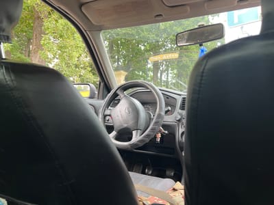 The image is taken from the back seat of a car, looking forward through the windshield. The interior of the car is visible, including the steering wheel, dashboard, and car seats. Outside the car, a tree-lined street is visible. A sign that reads "Парк Дубки" (Dubki Park) is prominently displayed in the distance. This suggests the car is located in or near Dubki Park in Kalininets, Russia. The environment appears to be a park or a street adjacent to a park, with lush green trees and foliage visible. The overall scene depicts a moment captured from within a vehicle, offering a glimpse of the surrounding environment and a local landmark.