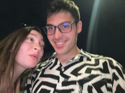 A selfie photo shows two people at night. A person with glasses is on the left, smiling. A second person with brown hair is on the right. Both are close to the camera. The person on the left is wearing a black and white patterned shirt. The background is dark, with a vertical light visible. The location is unidentifiable as Thessaloniki, Greece, based on visual information alone.