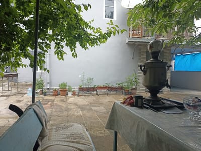 This image captures a quaint courtyard scene, likely in Gusar, Azerbaijan. In the foreground, a blue bench draped with a neutral-colored cloth is partially visible, suggesting a place for relaxation. To the right, a table covered with a plastic cloth is set up, with a decorative glass bowl filled with colorful items, possibly sweets or candies, and a samovar as the central object. The samovar, a traditional Russian and Azerbaijani tea urn, is made of brass and has a floral-patterned teapot on top. A small black box, perhaps containing tea, sits beside it.
Behind the table, a building with light grey walls and a white window is seen. Some greenery, including a tree with lush leaves, partially obscures the view of the building, creating a sense of an outdoor living space. Several potted plants are arranged in front of the building, adding to the garden-like atmosphere. A blue tarpaulin is visible in the background, suggesting some form of outdoor cover or shelter.
The overall impression is one of a peaceful, domestic setting. The lighting suggests it could be daytime, with overcast or diffused light, possibly indicating a cloudy day. The presence of the samovar hints at a moment of respite or a gathering for tea. There is no visible text in the image.