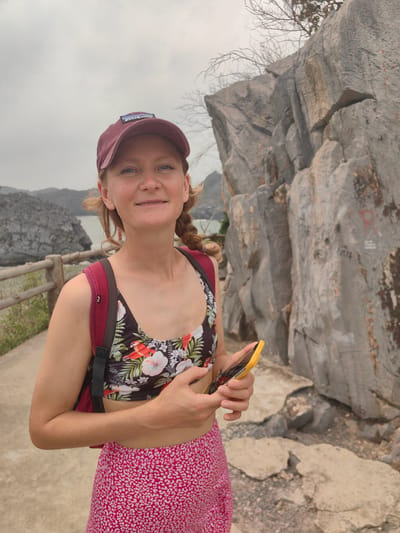 A woman stands on a concrete path holding a yellow smartphone. She is wearing a maroon Patagonia baseball cap, a floral-patterned sports bra, and a pink skirt with white patterns. A maroon backpack is visible on her shoulders. Behind her is a grey rock formation. In the background, there is a body of water and another rock formation, along with a wooden fence. The photograph was taken on Monkey Island, also known as Cat Ba Island, in the city of Hải Phòng, Vietnam.
