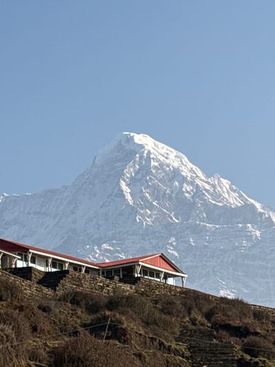 The image features a view of a mountain and architecture in Lwang Ghalel, Nepal. The sky is a clear blue, contrasting with a snow-covered mountain peak. The mountain appears to be a light gray color with a lot of texture throughout. A building is present on the right with a red roof. The building is dark and appears to be built into a hill.
