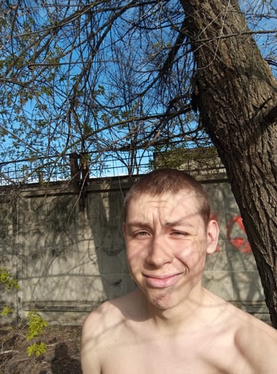 A selfie-style photograph shows a shirtless, light-skinned male with short brown hair, smiling with his head tilted to the left. The background reveals urban elements: a concrete wall with shadows and faded red graffiti, a metal fence, bare tree branches with some new leaves against a blue sky, and the textured bark of a tree trunk in the foreground. The image suggests an outdoor location in an urban setting. Given the specified location совхоза "Воронежский", Russia, it is assumed the image was taken there.