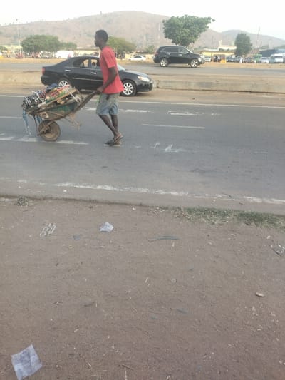 A man wearing a red t-shirt and blue jeans is pushing a wheelbarrow loaded with goods down a paved road in Maraba, Nigeria. He is walking to the right, away from the camera. The road is wide and has two white lines painted on it, dividing the lanes. There is a black car parked on the left side of the road, and a black SUV parked on the right side of the road. There are several other cars in the background, and buildings on the horizon. There are also some trees and bushes along the roadside. The ground is dirt and there is debris scattered on it. The sky is overcast.  The location is identified as Maraba, Nigeria based on the coordinates provided.