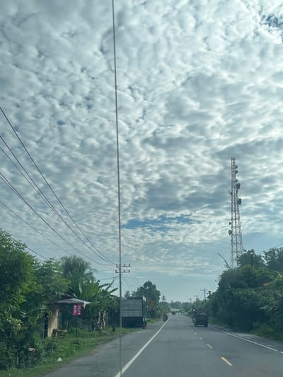 The image depicts a roadside scene in what appears to be Tu, Indonesia, under a sky filled with scattered, puffy clouds. A paved road runs through the center of the frame, with vehicles visible. On the left side of the road, there are several power lines stretching horizontally, a small wooden structure with a corrugated roof, dense green foliage, and banana trees. A large truck is parked or driving on the right side of the road. Further down the road, a tall telecommunications tower stands prominently against the cloudy sky. The environment suggests a rural or semi-urban setting with a mix of natural vegetation and infrastructure.