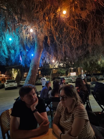 This image depicts a nighttime scene in Mitzpe Ramon, Israel. A group of people are seated at tables, likely at an outdoor restaurant or bar. The foreground features the top portion of a man with short, dark hair, wearing a black shirt and a watch. He is leaning on his hand. Another woman with glasses and a striped shirt is visible next to him. Further back, more people are seated, some looking up. The background contains a large tree illuminated with orange and blue lights. Cars are parked in the street behind the seating area. A sign is visible, but the text is not discernible. String lights illuminate a building further back.