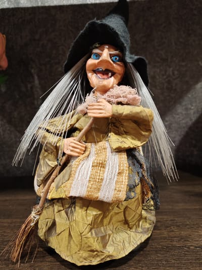 This is a full, front-facing studio portrait of a doll. The doll is dressed as a witch, wearing a black pointed hat and a gold, textured dress. The witch has bright blue eyes, a crooked nose, and a wide smile with visible teeth. Her hair is white and spiky, fanning out from her head. She is holding a small broom in front of her. The background is dark and textured, with a subtle hint of a pink flower on the left side of the frame. The doll is placed on a wooden surface. There is no indication of the city of Buy, Russia in the image.