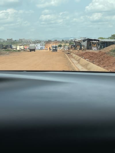 The image shows a view of a dusty, unpaved road in what appears to be Angwan Sayawa, Nigeria. The scene is captured from inside a vehicle, with a portion of the vehicle's interior visible on the right side of the frame. Along the left side of the road, there are makeshift structures and buildings of varying sizes, indicating a bustling commercial or residential area. A sign is visible, partially obscured, appearing to read "Welcome to." Several vehicles and motorcycles are present, along with people walking along the road. The sky is overcast with clouds. The landscape is somewhat uneven, with indications of construction and undeveloped areas along the edges of the road.