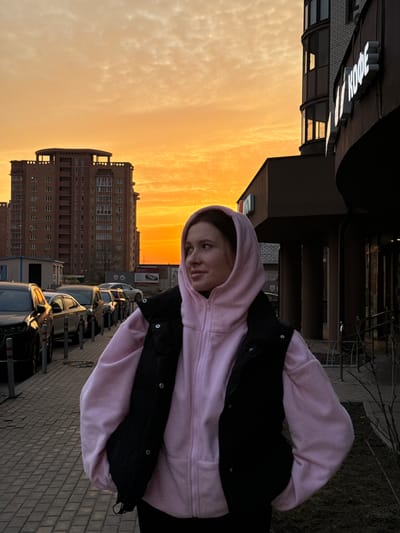 The image captures a female individual standing on a paved sidewalk in Gribovo, Russia, during what appears to be either sunrise or sunset, indicated by a sky transitioning from bright orange at the horizon to a lighter yellow-orange overhead, with scattered clouds. The female, positioned slightly to the right of the frame's center, is observed from the waist up. She wears a light pink zip-up hoodie with the hood covering her head, overlaid with a dark, possibly black, puffer vest. Her gaze is directed towards her right, looking away from the camera. To her left, a row of parked cars is visible along a curb, with the leftmost vehicle exhibiting front-end damage. Beyond the cars, a large multi-story residential building constructed of reddish-brown brick with numerous windows stands against the sky. To her right, a dark-colored commercial building with large windows features a white illuminated sign displaying the Cyrillic word "КОФЕ" (Coffee). The sidewalk is composed of gray pavers. Sparse, dark vegetation is present in the lower right foreground.