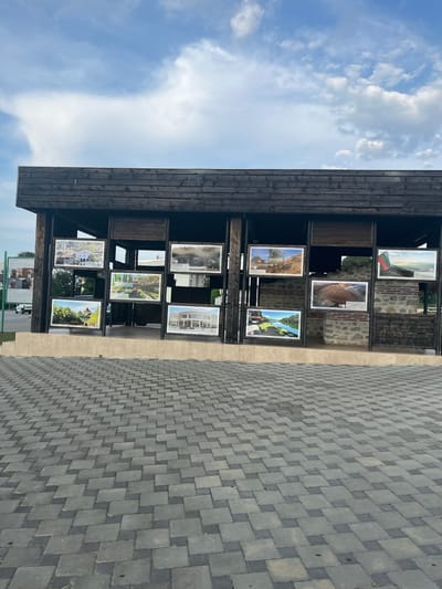 The image shows an outdoor exhibition of framed photographs displayed on a wooden structure. The photographs are arranged in a grid pattern, with some images visible on shelves. The structure itself is dark wood, and it is situated next to a paved area with a grey interlocking brick pattern. The sky above is blue with scattered white clouds, suggesting it is daytime.

The subjects of the photographs appear to be landscapes, architecture, and possibly historical scenes. One photo prominently displays the Bulgarian flag. Other images show rural or natural settings with buildings and greenery. One photograph appears to depict a modern building with large windows, possibly a museum or cultural center. Another shows a scenic view with a body of water and trees. Some of the photographs also contain text, which is too small to be legible in the provided image.

The scene appears to be a public space, likely an outdoor gallery or a part of a cultural attraction. The overall impression is one of an organized display of visual art in an open-air environment. The weather seems pleasant, suitable for an outdoor event. No people are clearly visible in the foreground or within the photographs themselves, focusing the attention on the displayed images and the setting. The location context provided is Petrich, Bulgaria, which suggests the exhibition may be related to the local heritage, culture, or natural beauty of the region.