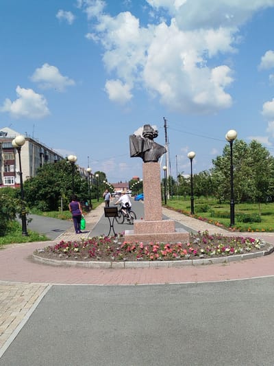 The image depicts a public space in the city of Yalutorovsk, Russia. In the foreground, a statue of a man's bust on a granite pedestal is centrally located within a flower bed adorned with colorful flowers. A paved walkway circles the flower bed, and beyond that, a road is visible. To the left of the statue, a woman in a purple shirt and dark pants walks away from the camera, carrying a green bag. Further in the background, a young person rides a bicycle on the paved walkway. Various streetlights with round, cream-colored lamps line the walkway. Behind the walkway and flower bed, there is a green lawn with trees, and residential buildings with balconies are visible on the far left. The sky is blue with scattered white clouds. The scene captures a bright, sunny day in a well-maintained urban park or public square.
