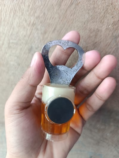 A hand is holding a novelty bottle opener designed to look like a miniature beer mug. The bottle opener, made of metal, is positioned at the top of the mug. The mug itself is made of clear glass and contains a golden-brown liquid, resembling beer, with a white frothy substance at the top. A circular black emblem is affixed to the side of the mug. The background is a wooden surface, possibly a table or countertop, with visible grain patterns. The image appears to be taken indoors, possibly in a home environment in Kawit, Philippines.
