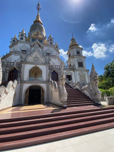 A low-angle, vertical shot of a white and gold temple with a brown stairway in Dĩ An, Vietnam. The temple is highly ornate with detailed carvings. A gold statue is visible through an archway. The staircase has serpent figures on either side. The sky is blue with white clouds. The sun is visible in the top left corner of the frame.
