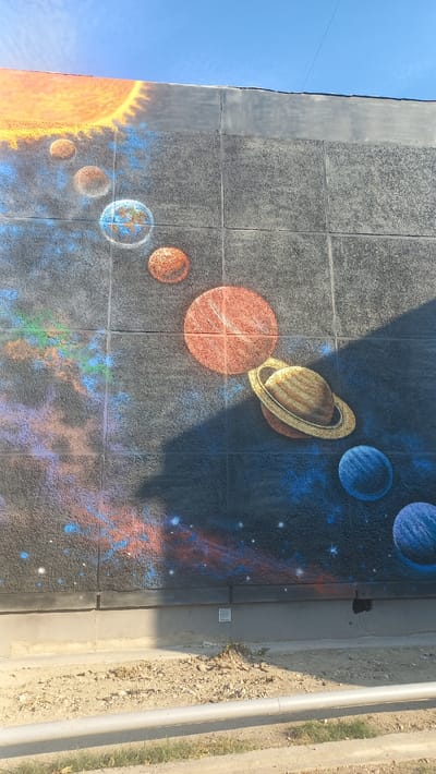 This media file displays a mural painted on a textured wall, depicting a solar system. The artwork features the sun on the left, rendered in vibrant oranges and yellows, surrounded by several planets. Among them are a ringed planet resembling Saturn, a large reddish-orange planet, and smaller, varied planets including one with blue continents and white clouds, suggesting Earth. The background is a deep blue speckled with white stars and nebulae painted in shades of red, blue, and purple, creating a cosmic scene. The mural is artfully painted, with shadows suggesting light sources and depth.

The mural is located on an exterior wall that appears to be part of a building. To the right of the mural, there is a concrete curb and a dirt/gravel area with some sparse green grass and a light-colored pipe running horizontally. The sky above the mural is clear and blue, indicating fair weather. The sun's position in the sky, casting shadows on the mural, suggests it is daytime. There are no people or specific activities depicted in the mural itself. The overall impression is one of artistic expression and educational display, likely representing the planets of our solar system. The texture of the wall, made of square panels, is visible beneath the painted imagery, adding a unique dimension to the artwork. No visible text is present in the image.