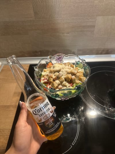 A person's hand with manicured fingernails holds an open bottle of Corona beer. In the background, a glass bowl filled with a salad topped with croutons and grated cheese sits on a black stovetop. The stovetop is situated in a kitchen with a wooden countertop visible on the left side. There is no indication of the city of Ruska Bela, Bulgaria.