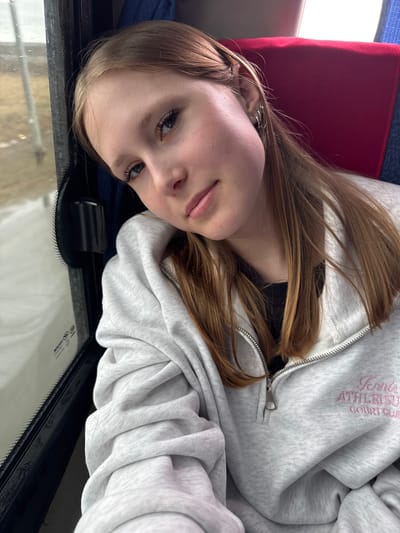 A young woman with light brown hair is positioned in what appears to be the interior of a vehicle or bus, evident by the seat and the window at the bottom of the image. She is wearing a light gray zippered hoodie with pink embroidery that reads "Tennis Athletics Court Club". Her head is tilted, and she looks directly at the camera. The vehicle's interior shows red and blue colored seating. The location is not discernible from the image, nor is there evidence provided that would place it specifically in Kukushtan, Russia.