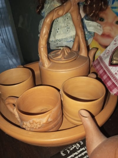 A terracotta tea set is displayed on a matching tray. The set includes a teapot with a sculpted handle and lid, accompanied by four cups. One cup has a floral design on its side. A doll with red hair is partially visible behind the tea set. An item with a red and white checkered pattern sits to the right of the teapot, and a black object is at the bottom edge of the picture.