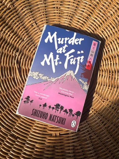 The scene is situated in Visp, Switzerland. A single paperback book, titled "Murder at Mt. Fuji" by Shizuko Natsuki, is positioned diagonally across a light brown, circular woven wicker surface. The book's cover features Mount Fuji with a snow-capped peak, set against a sky that transitions from dark blue at the top to vibrant pink and purple at the bottom. Silhouetted trees line the base of the mountain. Japanese characters, including "富士山殺人事件" (Fuji-san Satsujin Jiken - "Mount Fuji Murder Case"), are vertically oriented on the right side of the cover, partially obscured by a red, stylized blood splatter graphic. Review quotes from "TLS" and "Washington Post" are printed on the lower left and center-right of the cover, respectively. A Penguin Books logo is visible in the bottom right corner. Direct, strong lighting illuminates the book and the wicker, casting distinct shadows to the lower right, indicating a light source from the upper left. No individuals, actions, or interactions are visible.