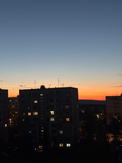 The image depicts a twilight scene over a residential area in the city of Neryungri, Russia. The sky transitions from a clear, deep blue at the top to a band of vibrant orange and red near the horizon, indicating the sunset. Silhouetted against the colorful sky are the upper floors of several apartment buildings. These buildings are of a typical Soviet-era design, characterized by their blocky structures and rows of windows. Many of these windows are illuminated, casting warm light that contrasts with the darkening surroundings. Antennas are visible on the rooftops of some of the buildings. The lower portions of the buildings and the foreground are almost entirely dark, with only faint outlines of trees and possibly parked cars visible. The overall impression is one of a quiet evening in a northern urban setting, with the last light of day providing a dramatic backdrop to the apartment blocks.