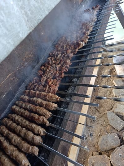 The image displays an outdoor cooking scene at Kasri Arifan, Uzbekistan, focusing on a long, dark metal grill. Approximately twenty-five metal skewers are laid across the grill's grates, actively cooking meat. In the foreground, ten skewers hold elongated, cylindrical ground meat kebabs, browned and appearing cooked. Further along the grill, fifteen skewers contain diced or cubed meat pieces, from which white-grey smoke actively rises, indicating cooking over hot embers or charcoal. The grill's interior is dark. To the left of the grill, a light-colored, textured wall is visible. To the right and in the immediate foreground, the ground consists of sandy soil mixed with irregular, broken stone or concrete pieces. No individuals are present in the scene.