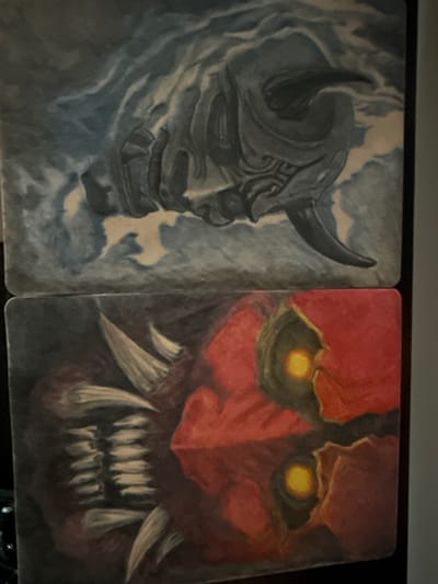 The image displays two distinct artworks stacked vertically. The upper artwork is a monochrome painting with shades of blue and grey, depicting a monstrous, horned head emerging from swirling clouds. The lower artwork is a vibrant painting featuring a creature with sharp teeth and glowing yellow eyes set against a predominantly red and black background. The overall style suggests a fantasy or horror theme. There is no visible indication of the location within the city of Leominster, United Kingdom.