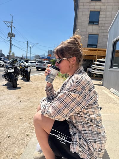 A woman is crouching on a paved surface in Toseong-myeon, South Korea, drinking from a paper cup. She wears sunglasses, a plaid shirt, and black shorts with white lettering down the side. Her hair is pulled back. Several motorcycles are parked along the street behind her. Cars are parked further down the street. Power lines are visible overhead, and buildings can be seen in the background.