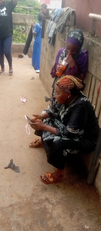 The image depicts a scene in what appears to be Idemili, Nigeria. Two women are prominently featured. One woman is seated, wearing a black outfit with silver embellishments and a beaded head covering, focused on a mobile phone she holds in her hands. A string of beads is also visible, dangling from the phone. She wears gold sandals. Beside her, slightly behind, another woman in a purple hijab is drinking from a bottle. In the background, other individuals are present: a person wearing a black shirt with text, jeans, and flip-flops; an individual in a blue garment; and another person further in the background. Clothes are draped over a metal barrier. The setting appears to be an outdoor concrete area next to a wall. On the ground, there is a small piece of trash and pink ribbon.