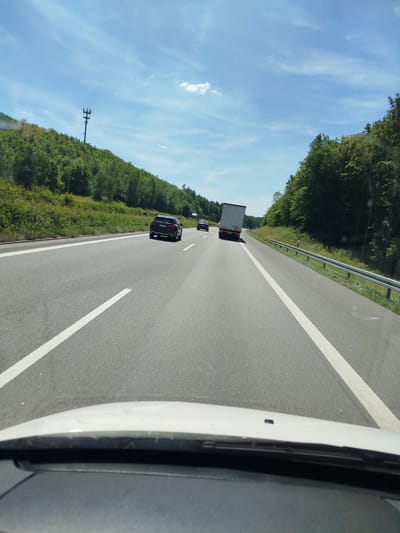 This is a forward-facing, eye-level shot from inside a vehicle, likely a car, traveling on a multi-lane highway. The road surface is asphalt with clear white lane markings. On the right side of the highway, there is a metal guardrail bordering a grassy shoulder and a dense forest of green trees. On the left side of the highway, the landscape consists of a steep, tree-covered hill with a tall cell tower visible near the crest.

Several vehicles are on the highway. Ahead and in the right lane, a dark-colored SUV is driving. In the left lane, further ahead, a white semi-trailer truck is visible, and behind it, a dark-colored passenger car. In the distance, on the left side of the highway, another dark-colored car is seen. The sky is bright blue with scattered white clouds, indicating daytime and fair weather. The lower portion of the image shows the dashboard and windshield of the vehicle from which the photo is taken, with some reflections visible on the windshield.

The location is identified as Estenfeld, Germany, a municipality in the Würzburg district.