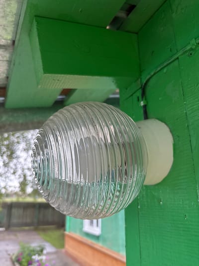 The image shows a close-up view of a light fixture mounted on a green wooden wall. The light fixture has a clear, ribbed glass cover with a white base. The glass cover is egg-shaped and appears to contain a bulb. The wooden wall is painted a vibrant green and shows some signs of wear, with visible grain and small imperfections. Above the light fixture, a green wooden beam is visible, also painted green. In the background, out of focus, is a blurry view of a yard with a wooden fence and some greenery. The overall impression is of an outdoor setting, possibly in a rural area. There is no indication of Gonja, Russia within the image itself.