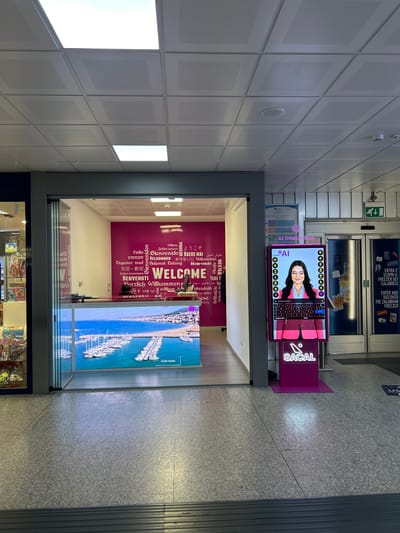 This media file captures an indoor scene, likely a public space such as an airport or a shopping mall, given the tiled walls, linoleum flooring, and the presence of retail displays and advertising. The image is oriented vertically.

In the foreground, a brightly lit retail space is visible, featuring shelves stocked with various products. The main subjects are two large digital displays. One display, prominently featured, shows a woman's face on an interactive screen with an "AI" logo and a keyboard interface, suggesting an AI-powered interactive kiosk. To the right of this, a large screen displays a vibrant image of a marina filled with white sailboats under a clear blue sky, with the text "Costa Ionica" superimposed.

Behind these displays, a wall is decorated with the word "WELCOME" in large, stylized letters, surrounded by the same word written in multiple languages, such as "Bienvenido," "Velkommen," and "Herzlich Willkommen," indicating an international arrival or greeting area.

Above and behind the interactive kiosk, a large advertisement promotes "THE CALABRIAN FREEZER" with Italian text that translates to "ENTER AND DISCOVER THE CALABRIAN FREEZER."

The lighting suggests an indoor environment, possibly daytime due to the bright illumination. The overall impression is one of modern infrastructure designed to welcome and engage visitors, with a focus on technology and international greetings. The presence of multilingual text and the marina imagery hints at a connection to travel or tourism, potentially related to the Calabria region of Italy, as suggested by the advertisement.