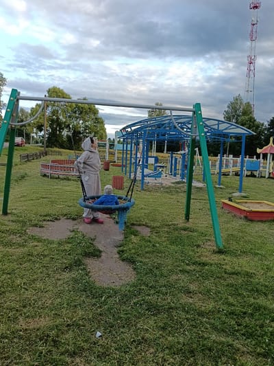 In a park setting in Smolino, Russia, a woman stands near a swing set, looking up at the sky. Her companion, a baby, sits in a circular swing designed for young children. The woman is wearing a light-colored hooded tracksuit and pink shoes, while the baby is dressed in a blue outfit and hat. The playground features various equipment, including a blue metal structure with a roof, a sandbox with red and yellow borders, and a circular red bench. In the background, trees and a tall communication tower are visible. The sky is filled with clouds, suggesting it might be late afternoon or early evening. The overall atmosphere appears peaceful, with the woman seeming to contemplate the sky as she supervises the baby. No visible text is present in the image.