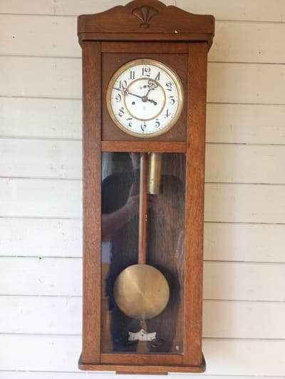 A full-shot medium of a wooden grandfather clock with a white face and black numerals, hanging on a white wooden wall. The clock has a round, gold-colored pendulum and a glass door that reveals a reflection of a person. The clock's hands are pointing to 10:10. The clock is positioned centrally in the frame, and the wooden wall behind it is made of horizontal planks. The overall lighting suggests it is daytime. The scene is indoors or on a covered porch, as indicated by the wall construction. The setting appears to be a rustic or traditional environment. There is no visible text in the image.